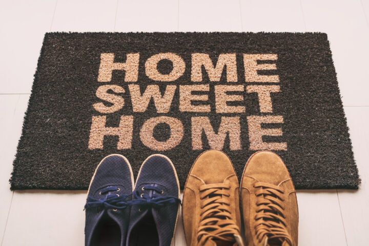Door mat Shoes at front entrance of condo apartment. Written welcome sign Home Sweet Home welcoming homeowners at new house moving in couple's pairs of sneakers lying on the floor.