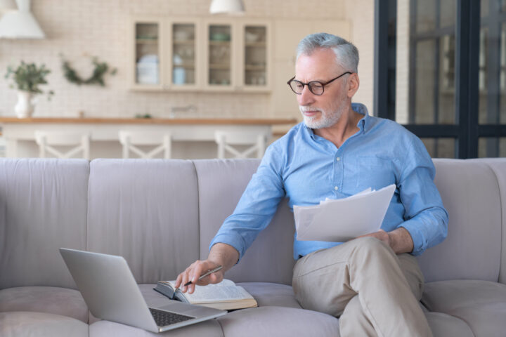 Working serious old senior elderly businessman paying domestic bills online using laptop, counting, paperwork, home office. Social distancing concept