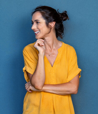 Mature beautiful latin woman in yellow dress. Portrait of positive brunette woman smiling and looking away. Happy middle aged lady standing against grey wall and thinking.