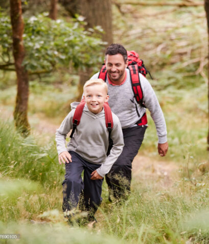 A boy and his father walking together on a trail between trees in a forest, both smiling, elevated view