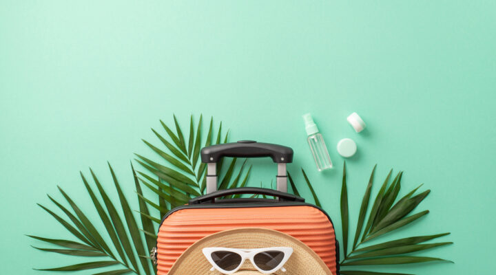 A captivating turquoise setting sets the stage for a top view of a suitcase, beach gear, sunglasses, headwear, spf cosmetic bottles on palm leaves. Great for travel marketing